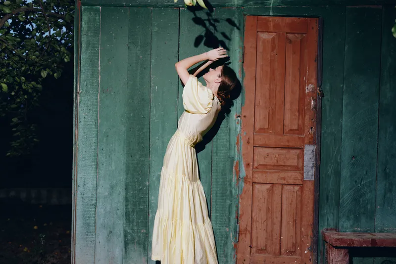 Young woman in a pale yellow dress leans against a green wall beside a wooden door, captured in soft daylight with a graceful vintage vibe.