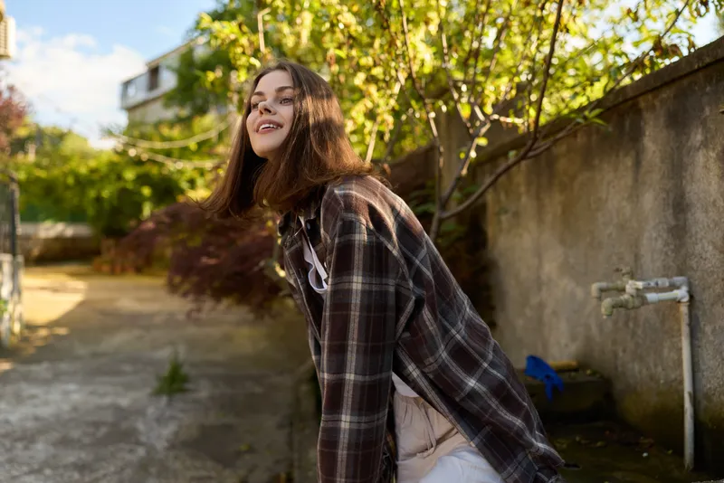 Joyful woman enjoying outdoor lifestyle moment, wearing plaid shirt, casual attire, sunlit garden scene, candid smile, relaxed pose, natural light, everyday fashion, fresh air, carefree mood