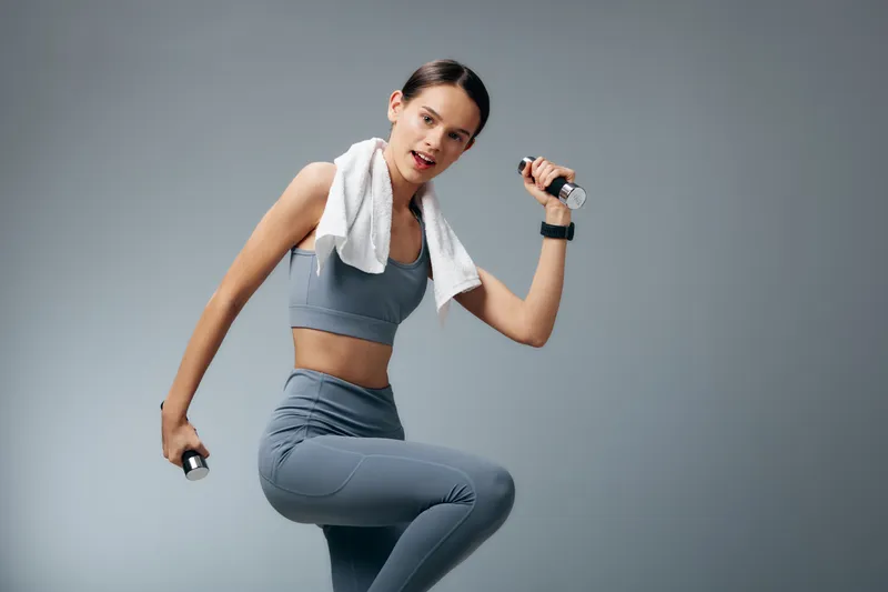 Fitness-focused woman in seamless athletic wear performs a dynamic workout with dumbbells, towel over shoulder, against a plain studio backdrop, showcasing strength and movement.