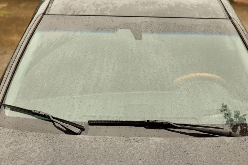 Rain smeared windshield of a car with droplets on wet glass, wipers at rest, window and exterior reflecting surroundings, conveying weather, travel, and outdoor routine in the morning light