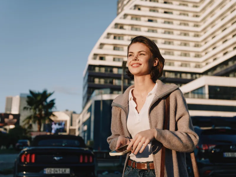 A cheerful business professional is seen texting outdoors near a modern building. She smiles and appears engaged, creating a positive and dynamic work atmosphere.