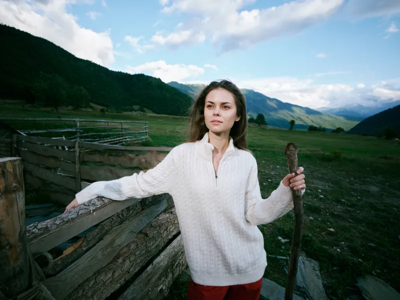 Woman outdoors by a wooden fence in a vast rural landscape holding a staff with mountains and open fields behind, wearing a light sweater under a bright cloudy sky