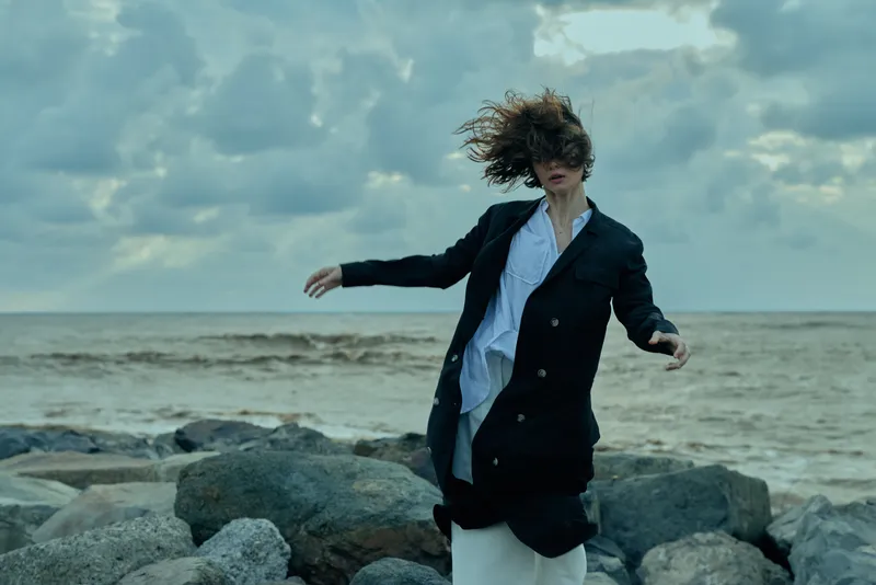 Windy beach motion with a long coat flapping as a figure stands on rocks by the sea, dramatic sky and outdoor fashion on the shoreline