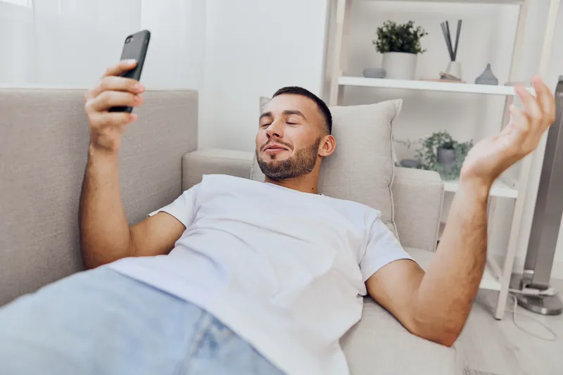 Relaxed man enjoying his smartphone on a cozy sofa, portraying modern living and digital connectivity in a serene environment, perfect for lifestyle themes.