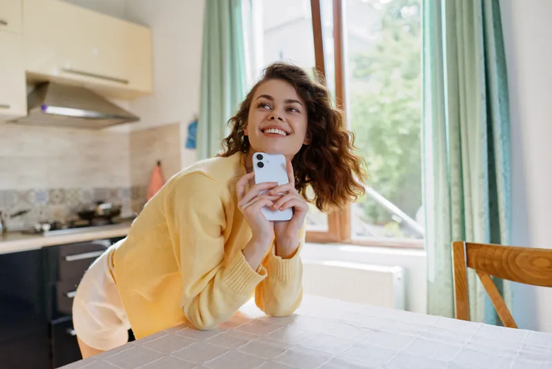 Young woman in a yellow sweater leans on a kitchen table, smiling and looking at a smartphone in a candid moment of everyday life.