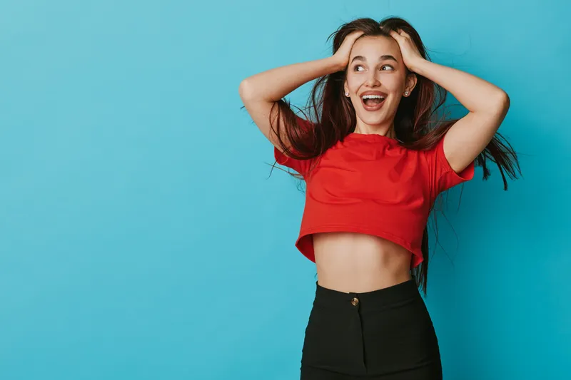 Young woman with long hair wearing red crop top and black pants posing with hands on head and smiling against blue background in studio for lifestyle portrait.