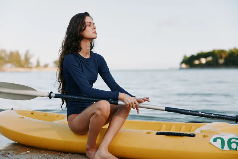 A woman sits in a yellow kayak at the shoreline, hands resting on a paddle, calmly gazing toward the sea, with soft light and a tranquil summer mood.