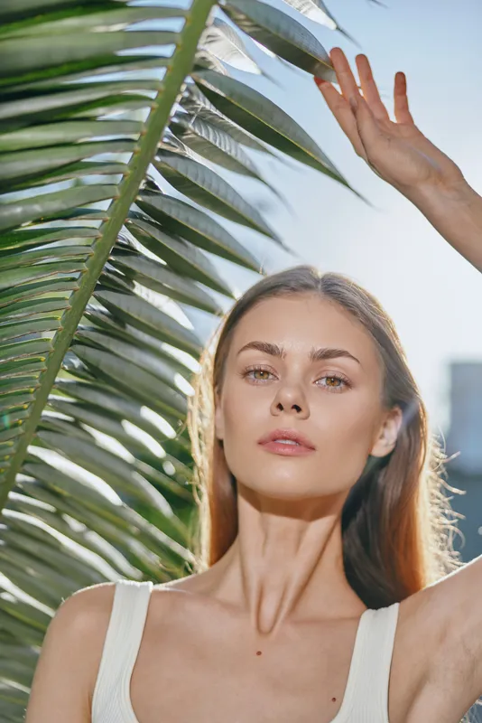 Young woman with natural skin and light hair stands outdoors under bright sunlight holding a palm leaf, wearing a white sleeveless top in a serene summer setting.