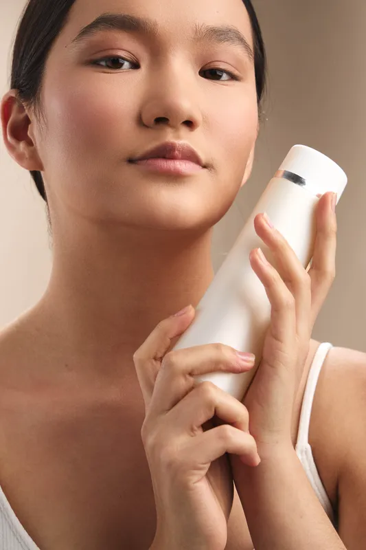 Asian woman holds a white bottle close to her face, showcasing a skincare routine in a calm home setting. The composition conveys beauty, care, and everyday wellness.