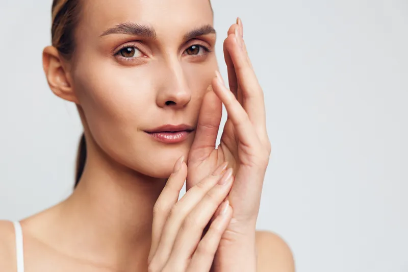 Closeup portrait of young woman with clear skin touching her face gently, looking at camera with calm expression, isolated on light background in skincare concept.