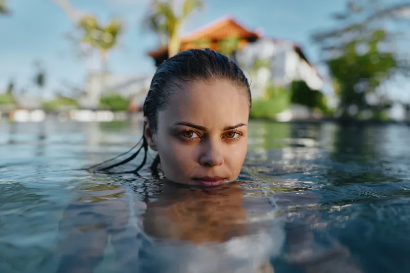 Young woman with wet hair swims calmly in a clear outdoor pool on a sunny day, showing a relaxed and focused expression with blurred background.