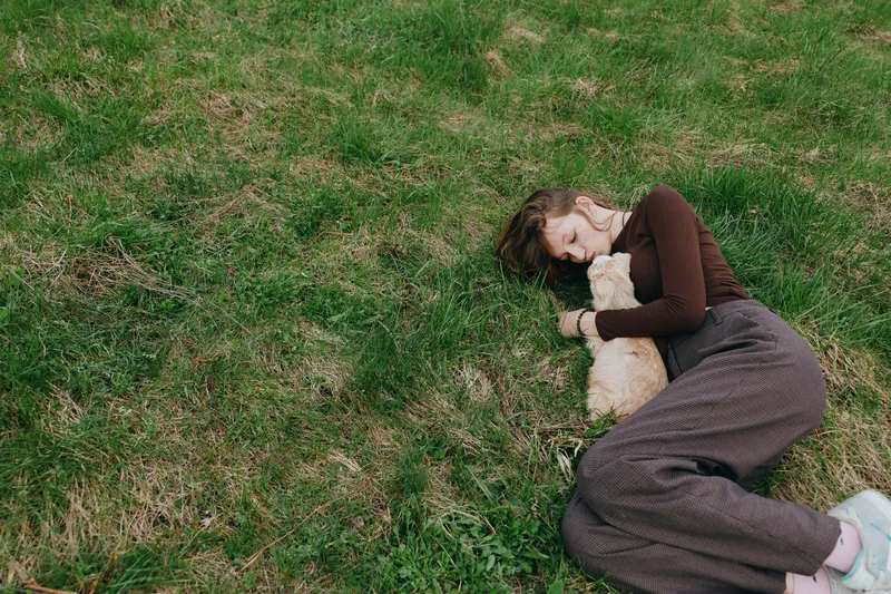 Young woman lies on green grass holding a small goat close to her chest in a peaceful outdoor lifestyle moment during springtime.