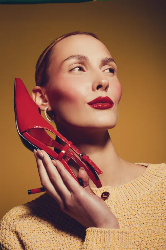 Woman with red lipstick holds red high heel shoe near face, wearing yellow sweater. Studio portrait on yellow background. Concept of fashion, style, and beauty accessories.