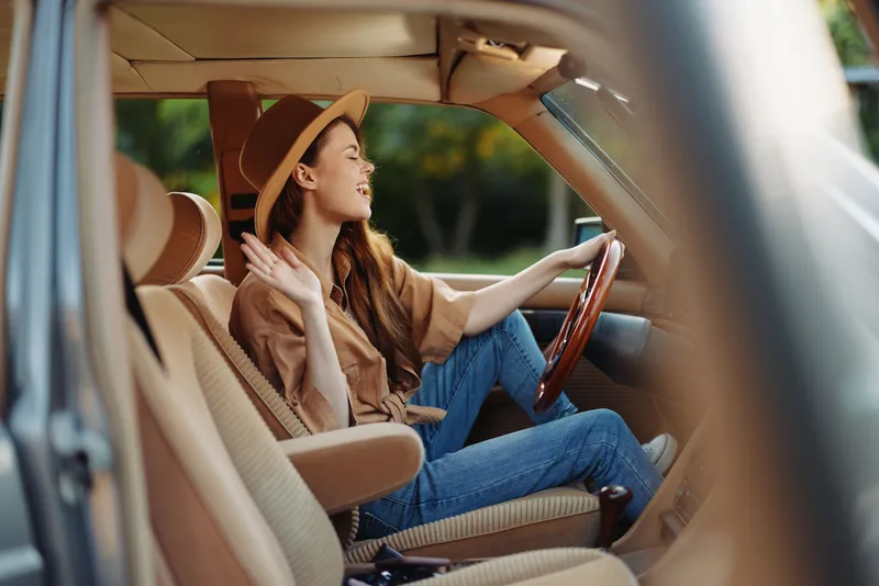 Cheerful young woman in stylish outfit enjoying a drive in a vintage car, wearing a hat and smiling with a warm, sunny background.