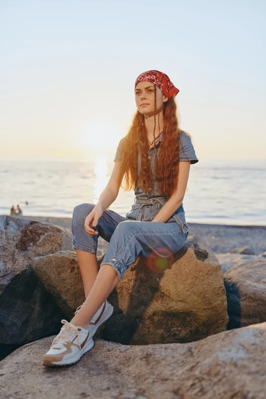 Lifestyle woman in modern boho-western denim street style sits on rocks by the sea during golden hour with a red bandana and casual sneakers, film color effect.