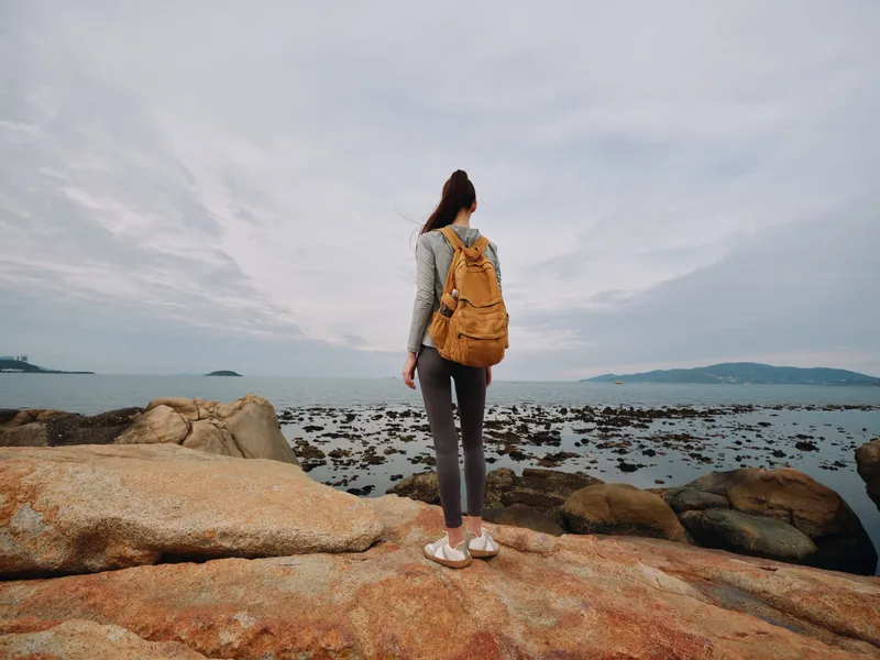 Young woman in casual outfit with a backpack, standing on rocks near the seaside, enjoying a calm day at the beach