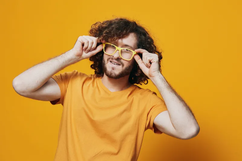 Cheerful young man with curly hair wearing yellow glasses and a bright yellow t shirt against a vibrant orange background, exuding a playful and trendy vibe