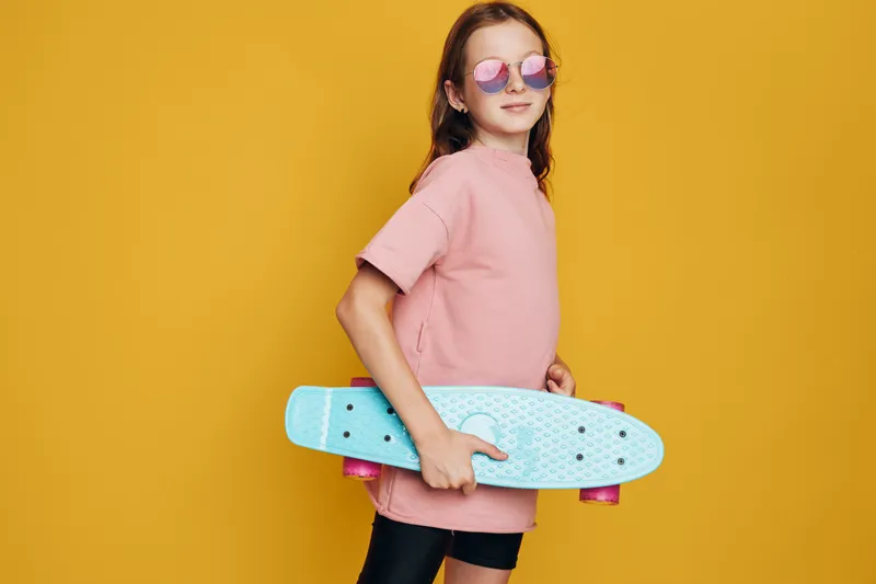 Girl in pink shirt holds blue skateboard confidently wearing round sunglasses on yellow background.