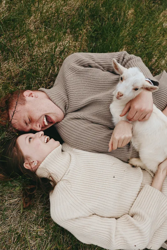 goat, kid, couple, outdoors, grass, affection, warmth, cuddle, nature A couple lie in cozy sweaters on grass, smiling while cradling a playful goat kid, illustrating rural lifestyle, connection, and