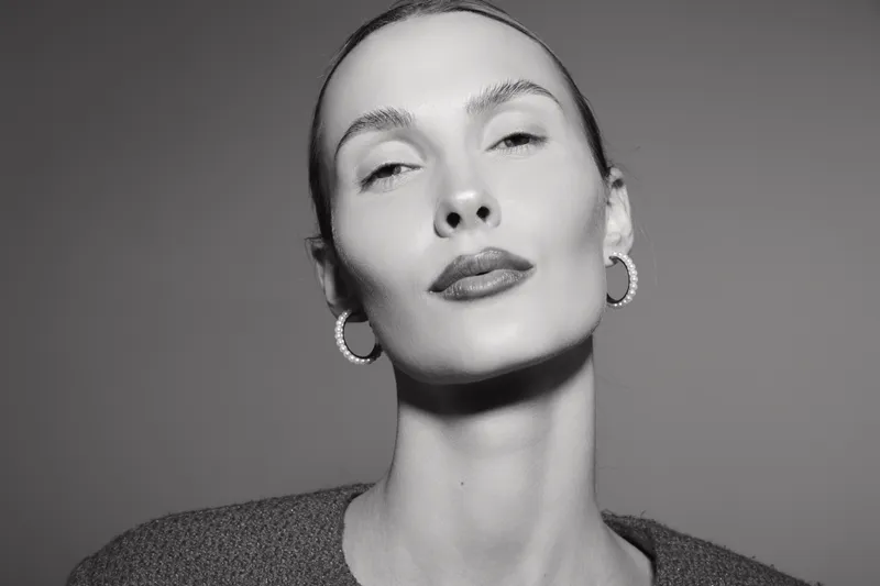 Close-up of a pale Caucasian woman with slicked back hair and hoop earrings posing confidently in black and white studio lighting.