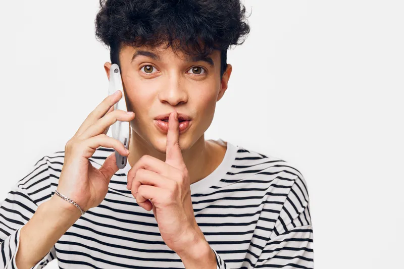 Young man in striped shirt making a shushing gesture while talking on the phone, expressing secrecy and quiet communication in a bright studio setting.