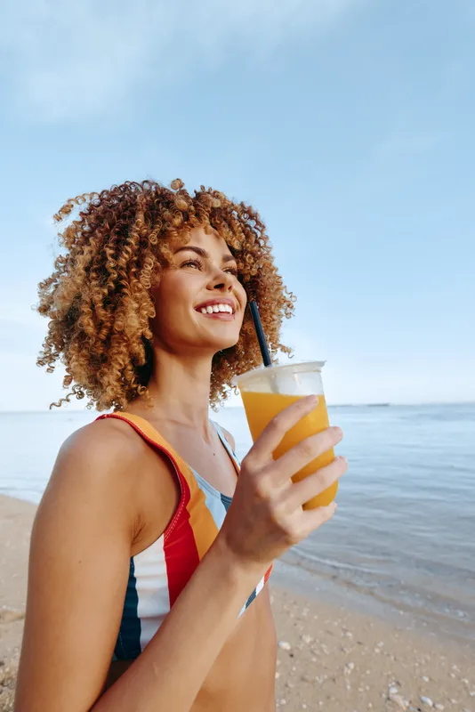 Cheerful woman on a sunny beach holds a glass of juice, smiling at the camera, enjoying a summer vacation by the ocean under a bright blue sky, relaxed outdoor mood