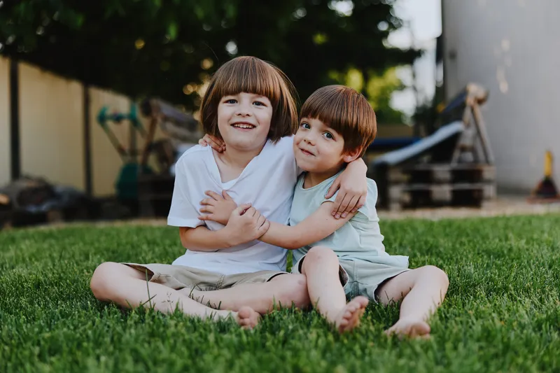 Two joyful boys sitting together on green grass, enjoying a sunny day outdoors with smiles and affectionate hugs that convey warmth and friendship.