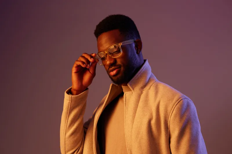 Stylish young Black man wearing glasses and a trendy coat, expressing confidence against a gradient purple backdrop in a modern indoor setting