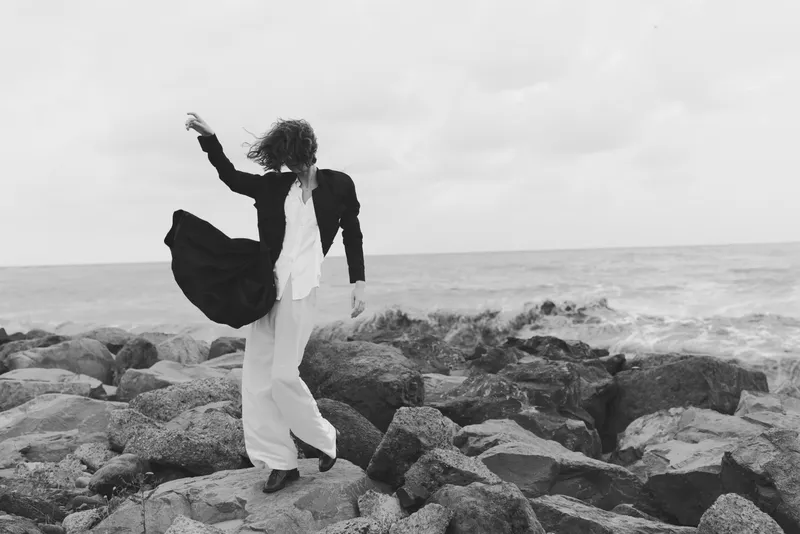 Monochrome scene of a stylish figure standing on rugged rocks beside the sea, coat billowing, white pants, wind swept hair, exploring freedom and bold movement on a dramatic coastline.