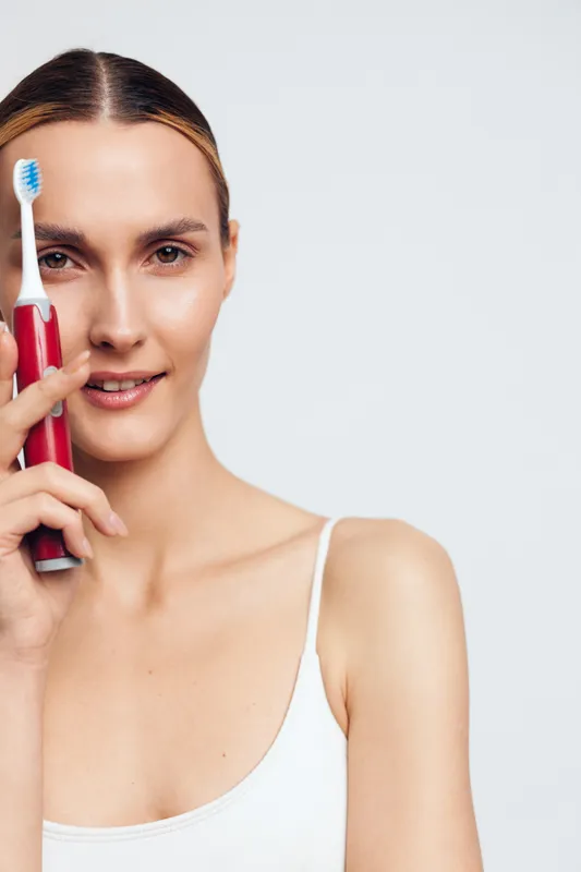 Young woman holds a toothbrush near her face, smiles at the camera, clean skin, white top, studio light, minimalist portrait focusing on dental care and personal hygiene.