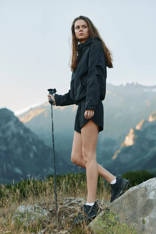 A solitary hiker stands on a rocky trail in a rugged mountain landscape. She wears a dark windbreaker, shorts, and sturdy boots, poised and focused on the distant path ahead.