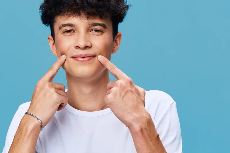 smiling man in white t-shirt pointing at his cheeks with fingers against blue background, young adult, casual style, joyful expression, studio portrait