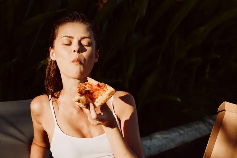 Smiling woman in a white top eats a slice of pizza outdoors, enjoying a summer casual lifestyle moment with food, snack, sunlight, warm atmosphere and relaxed mood.
