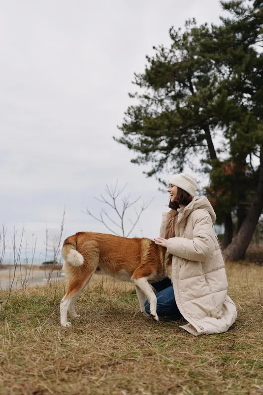 Woman kneeling on a grassy meadow outdoors, petting a dog in a warm winter coat, with trees and a calm landscape, capturing a peaceful moment of companionship