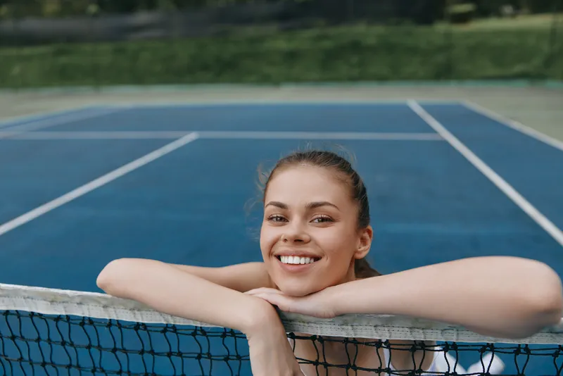 Young female tennis player smiling on the court, showcasing sports enthusiasm and joy in a vibrant blue setting, perfect for sports and wellness concepts