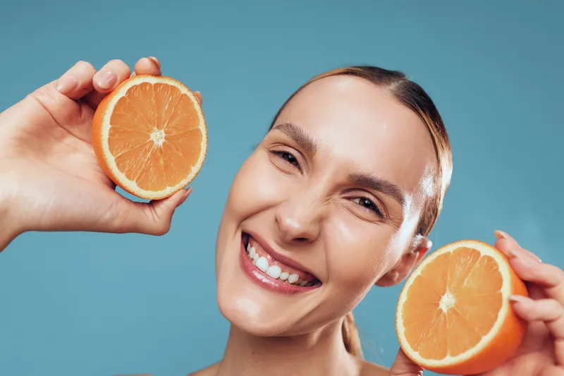 Smiling woman holding two halves of fresh orange fruit close to her face, bright cheerful expression, healthy skin, clean background, natural vitamin C concept