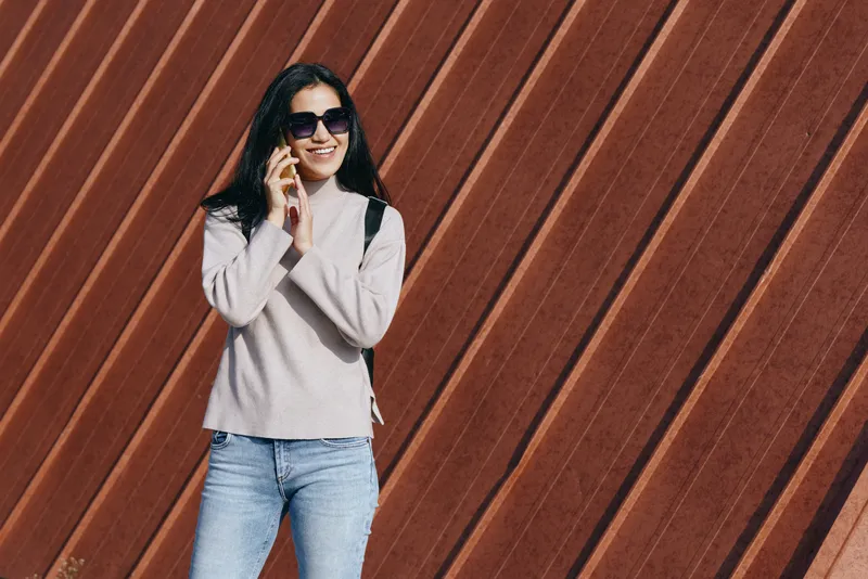 Woman wearing sunglasses, casual sweater and jeans, leaning on a diagonal wooden wall, talking on a phone, showcasing urban lifestyle and stylish confidence