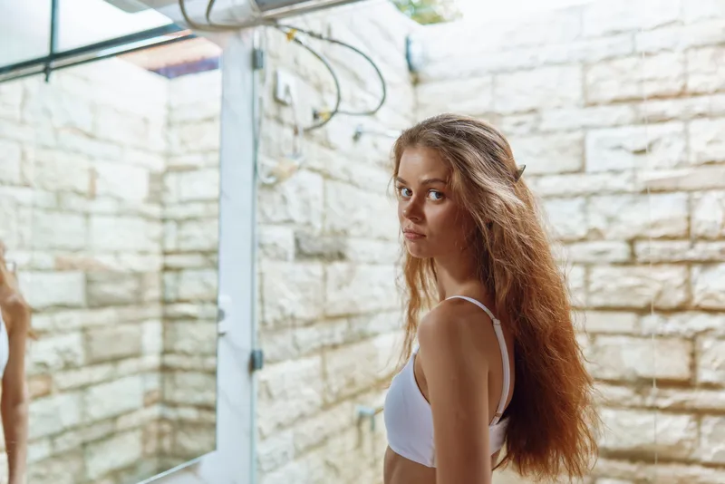 Woman with long hair wearing white top standing near rustic stone wall in soft natural light, looking contemplative with neutral expression indoors.