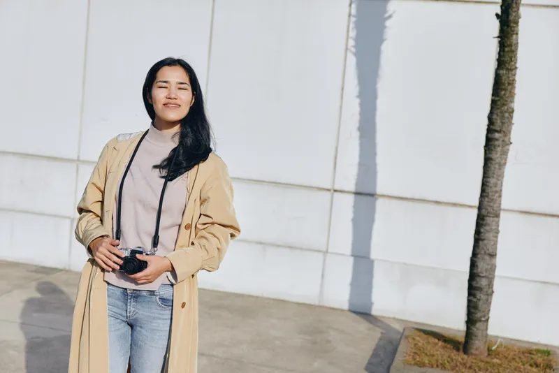 Woman photographer standing outdoors with camera in hand, wearing a beige coat and denim jeans, sunny day, urban backdrop, relaxed pose capturing candid street moments for street photography