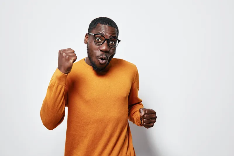 African American man with dark skin wearing glasses and orange sweater showing excitement and celebration with clenched fists on white background. Studio portrait of happy male.
