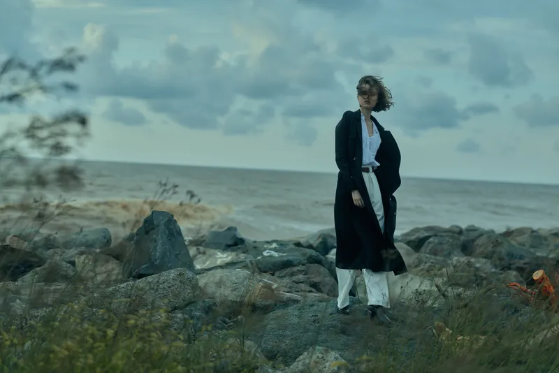 Woman in a long coat stands on rocky shore with the sea behind, windy day outdoor fashion scene capturing movement and rugged coastal mood