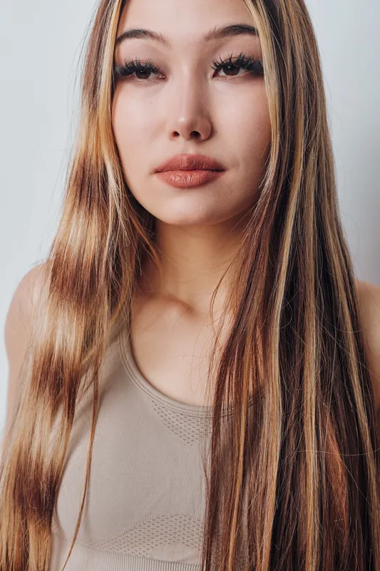 Young woman with long straight hair and natural makeup posing indoors wearing a casual sleeveless top. Beauty portrait with smooth skin and neutral expression on light background.