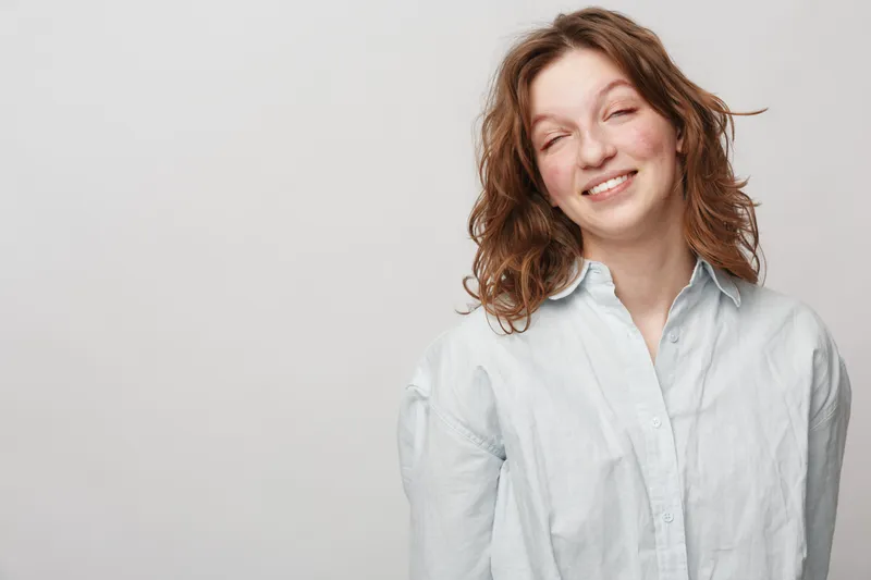Smiling young woman with wavy brown hair wearing white shirt stands against light background. Natural expression and casual style create relaxed and friendly portrait concept.