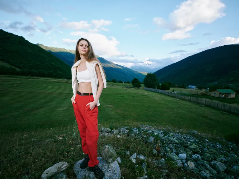 Woman standing on rocky ledge in a green valley with distant mountains, casual white crop top and red pants, expansive landscape, nature and tranquil outdoors scene