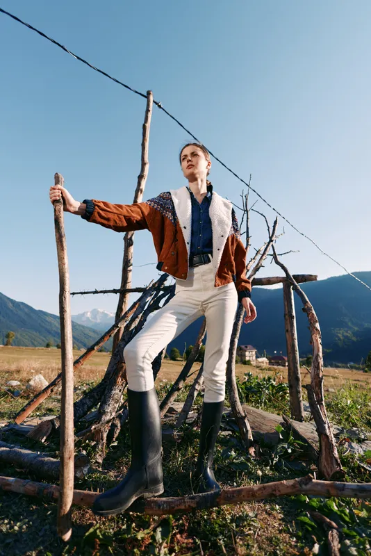 Woman in jacket and hiking boots standing by a wood fence in the countryside mountains, rural nature scene with clear sky, confident pose and outdoor adventure atmosphere.