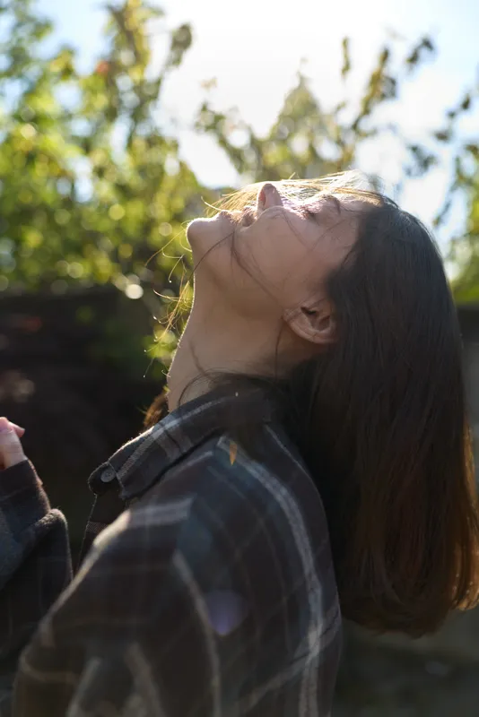 Woman outdoors daylight sunlight portrait lifestyle, a plaid shirt casual scene with a serene mood, head tilted back and eyes closed in a bright natural setting, calm and content