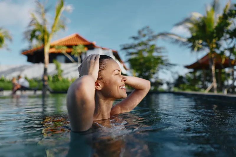 A poolside scene shows a woman in clear water, enjoying relaxation and vacation vibes at a resort, with summer sunlight, a carefree smile, and happiness radiating as she lounges and swims.