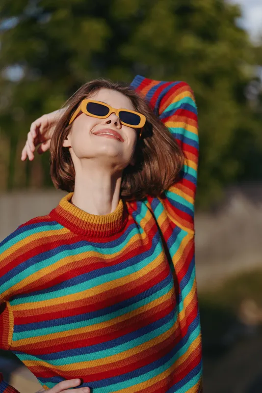 Woman in a colorful striped sweater wearing orange sunglasses, smiling with short hair outdoors in warm sunlight, casual outfit and confident pose for lifestyle, fashion or travel imagery.