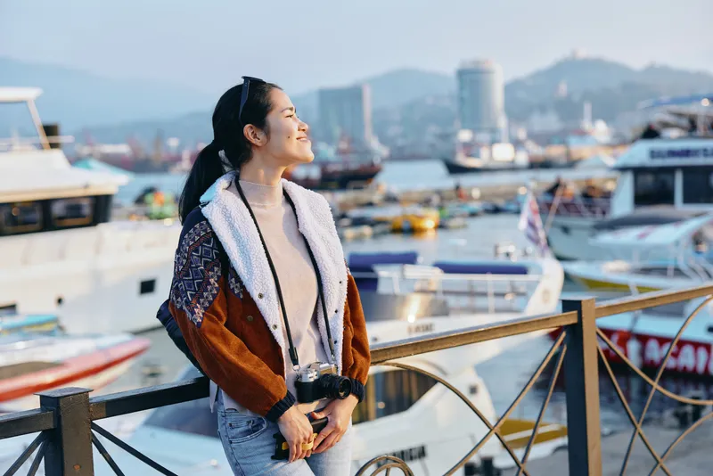 A woman in a warm scarf and patterned coat stands by a marina, gazing toward the harbor with a bright smile as boats rest on calm water and a distant cityscape frames the scene.