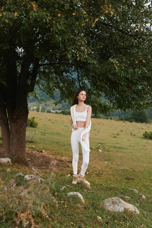 Confident woman in a white outfit posing outdoors under a large tree, relaxed stance, sunlit countryside, natural colors, peaceful mood, nature backdrop, casual style